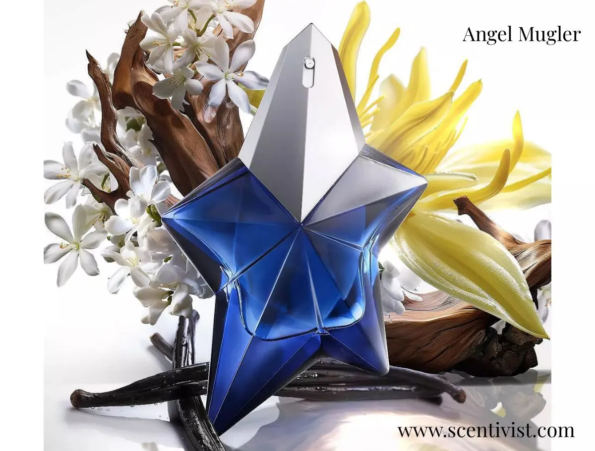 A blue, star-shaped Angel Mugler perfume bottle surrounded by white blossoms, vanilla pods, and warm woody elements on a soft, light background.