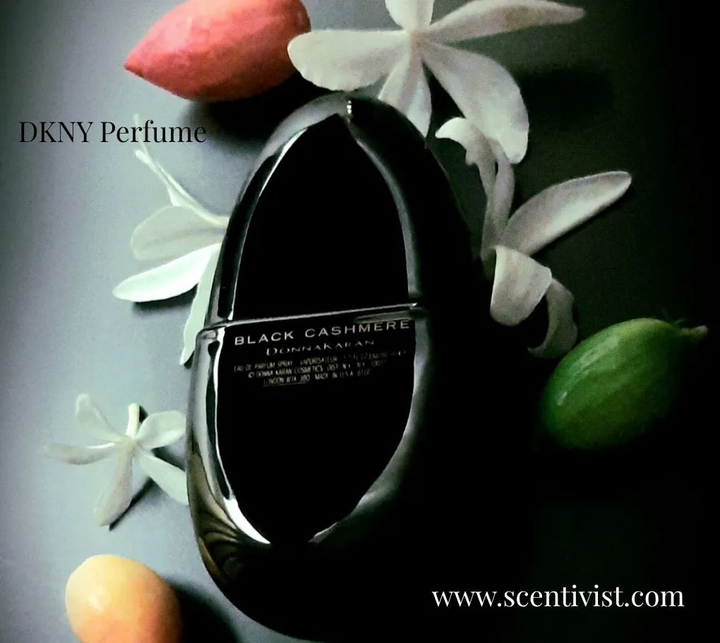 DKNY Black Cashmere perfume bottle surrounded by white flowers and colorful fruits on a soft, moody background.